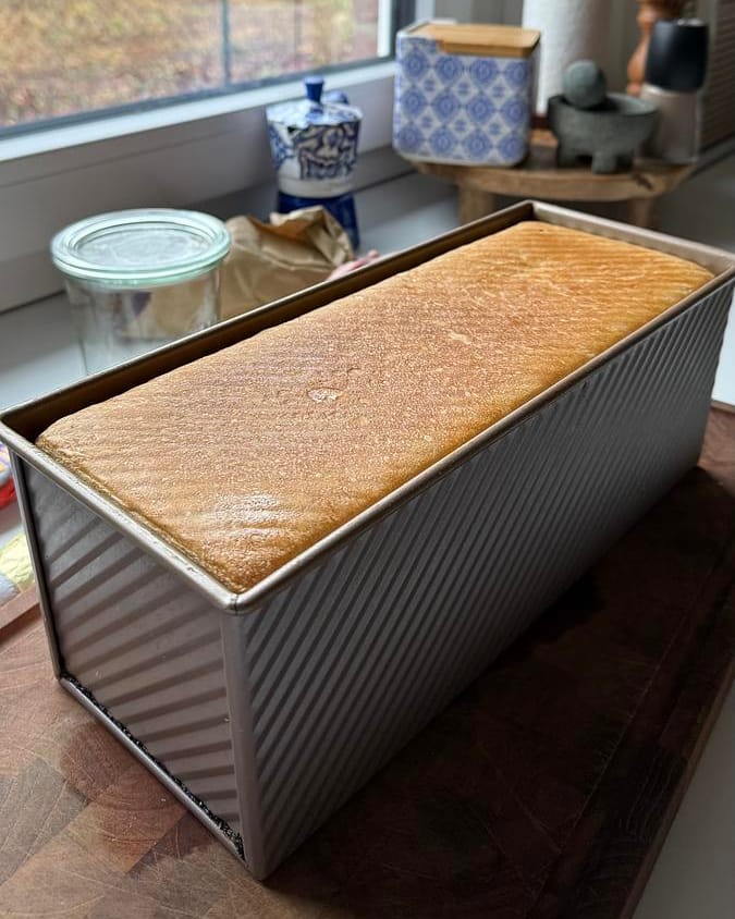A baked sandwich loaf, golden on top, in its pan with the lid off